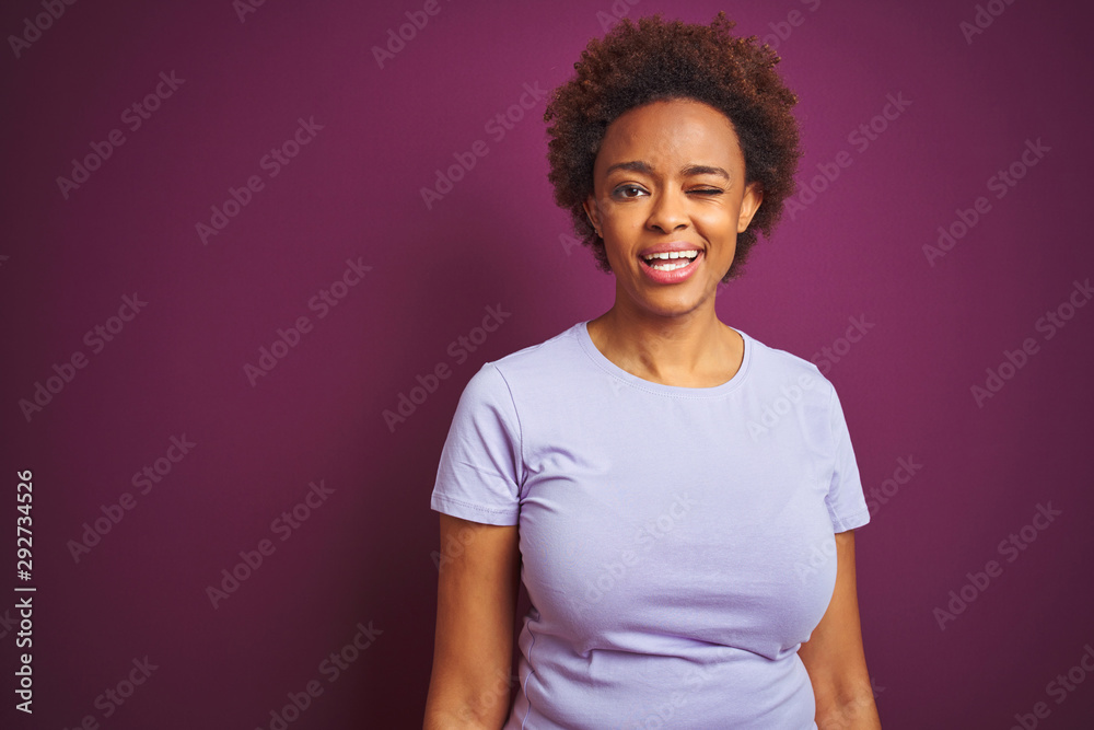Obraz premium Young beautiful african american woman with afro hair over isolated purple background winking looking at the camera with sexy expression, cheerful and happy face.