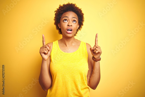 Beauitul african american woman wearing summer t-shirt over isolated yellow background amazed and surprised looking up and pointing with fingers and raised arms.