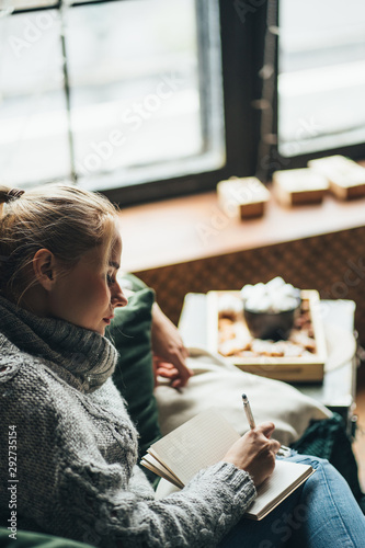 Christmas wish list. Woman relaxing at coach, eating sweets, making notes in book. Lazy weekend, cozy atmosphere, winter holidays, hygge concept