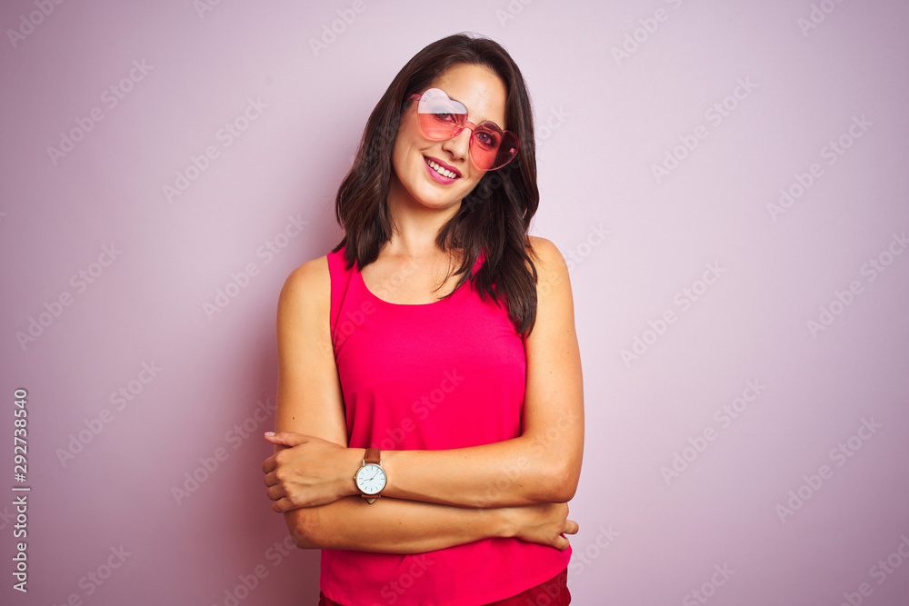 Beautiful woman wearing funny pink heart shaped sunglasses over pink isolated background happy face smiling with crossed arms looking at the camera. Positive person. Beautiful woman wearing funny pink heart shaped sunglasses over pink isolated background happy face smiling with crossed arms looking at the camera. Positive person.