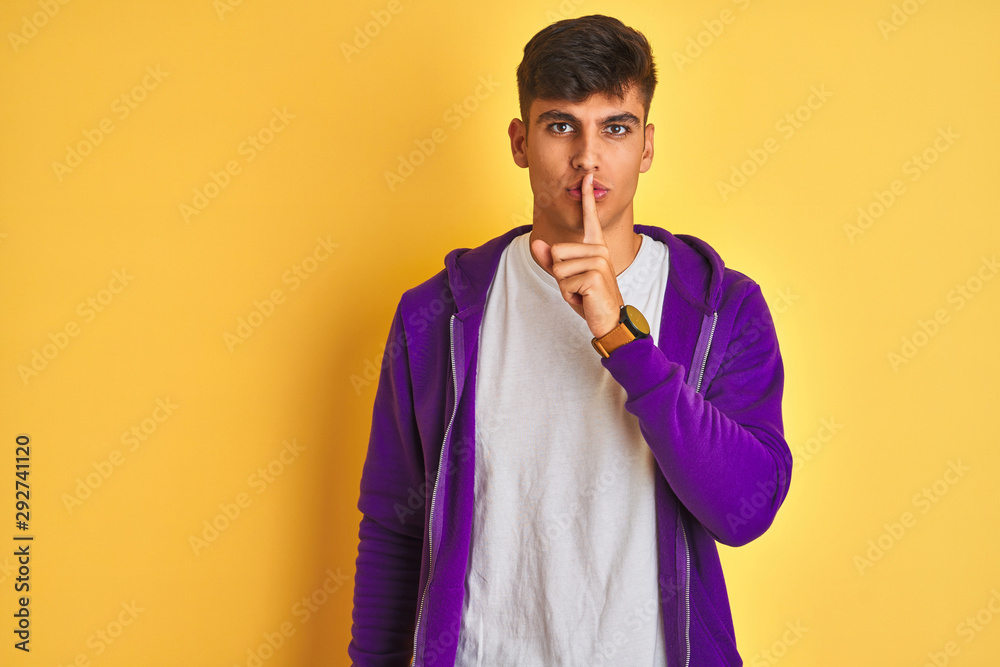 Young indian man wearing purple sweatshirt standing over isolated yellow background asking to be quiet with finger on lips. Silence and secret concept. Young indian man wearing purple sweatshirt standing over isolated yellow background asking to be quiet with finger on lips. Silence and secret concept.