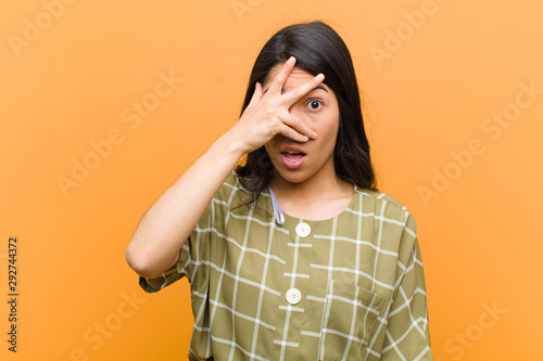 young pretty hispanic woman looking shocked, scared or terrified, covering face with hand and peeking between fingers against brown wall photo