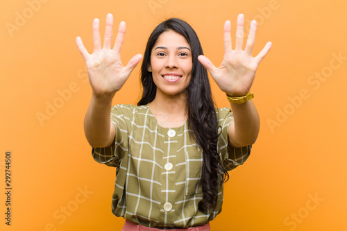 young pretty hispanic woman smiling and looking friendly, showing number ten or tenth with hand forward, counting down against brown wall photo