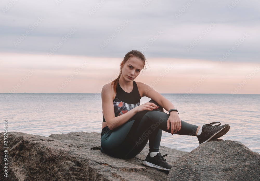1 white European young woman in sportswear sitting on a rock by the sea at sunset