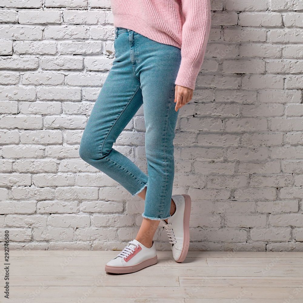 Female legs in turquoise jeans, pink sweater and sneakers leaning to white brick wall