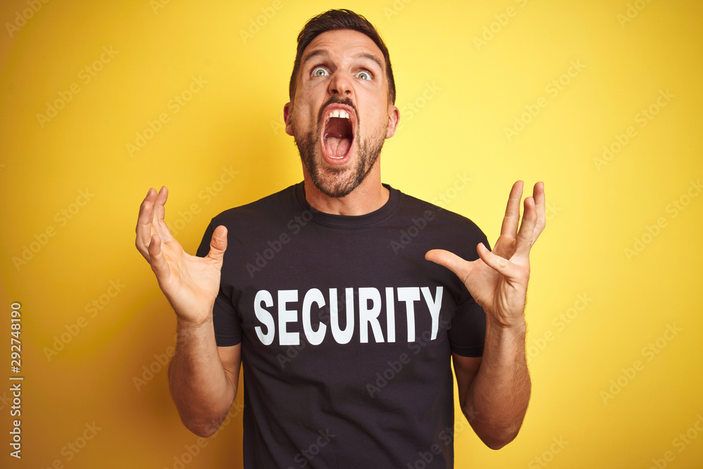 Young safeguard man wearing security uniform over yellow isolated background crazy and mad shouting and yelling with aggressive expression and arms raised. Frustration concept. Young safeguard man wearing security uniform over yellow isolated background crazy and mad shouting and yelling with aggressive expression and arms raised. Frustration concept.
