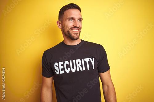 Young safeguard man wearing security uniform over yellow isolated background looking away to side with smile on face, natural expression. Laughing confident.