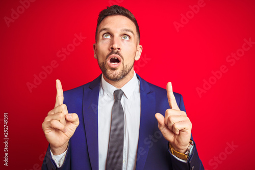 Young handsome business man over red isolated background amazed and surprised looking up and pointing with fingers and raised arms.