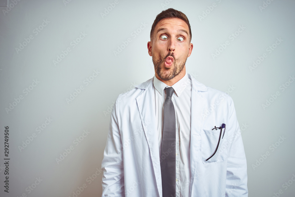 Young handsome doctor man wearing white profressional coat over isolated background making fish face with lips, crazy and comical gesture. Funny expression. Young handsome doctor man wearing white profressional coat over isolated background making fish face with lips, crazy and comical gesture. Funny expression.