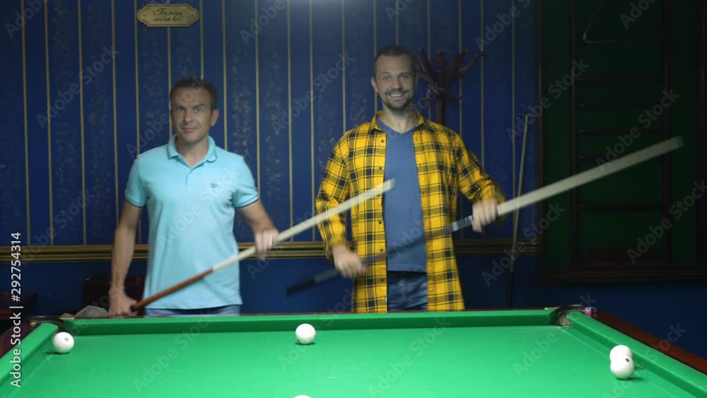 two men are dancing with a cue in their hands while standing next to the pool table. Fun with friends while playing billiards.