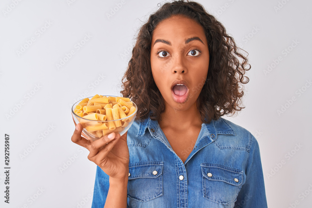 Young brazilian woman holding bowl with macaroni pasta over isolated white background scared in shock with a surprise face, afraid and excited with fear expression Young brazilian woman holding bowl with macaroni pasta over isolated white background scared in shock with a surprise face, afraid and excited with fear expression