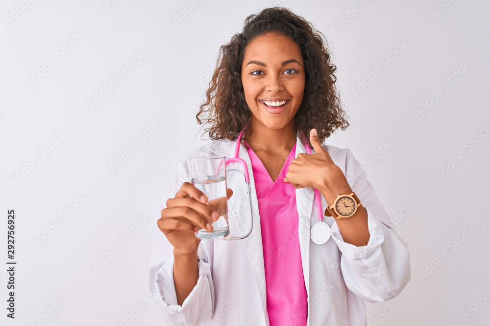 Young brazilian doctor woman holding glass of water standing over isolated white background with surprise face pointing finger to himself Young brazilian doctor woman holding glass of water standing over isolated white background with surprise face pointing finger to himself