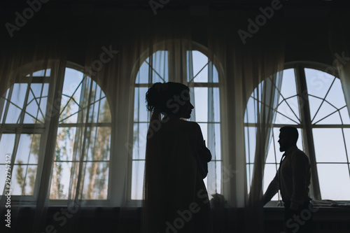 Stylish groom and beautiful bride stand near a large window. Wedding portrait of the newlyweds in the room, studio. Silhouette, photography and concept, profile.