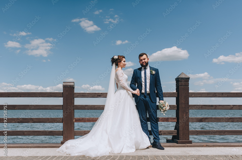 Beautiful newlyweds are standing on a wooden bridge, against the background of the sky and the sea. Classic wedding portrait of a stylish bride and groom. Photography and concept. Beautiful newlyweds are standing on a wooden bridge, against the background of the sky and the sea. Classic wedding portrait of a stylish bride and groom. Photography and concept.