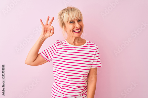 Middle age woman wearing striped t-shirt standing over isolated pink background smiling positive doing ok sign with hand and fingers. Successful expression.