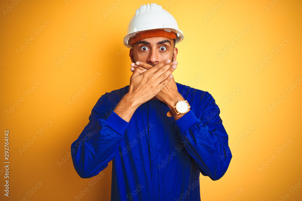 Handsome indian worker man wearing uniform and helmet over isolated yellow background shocked covering mouth with hands for mistake. Secret concept.