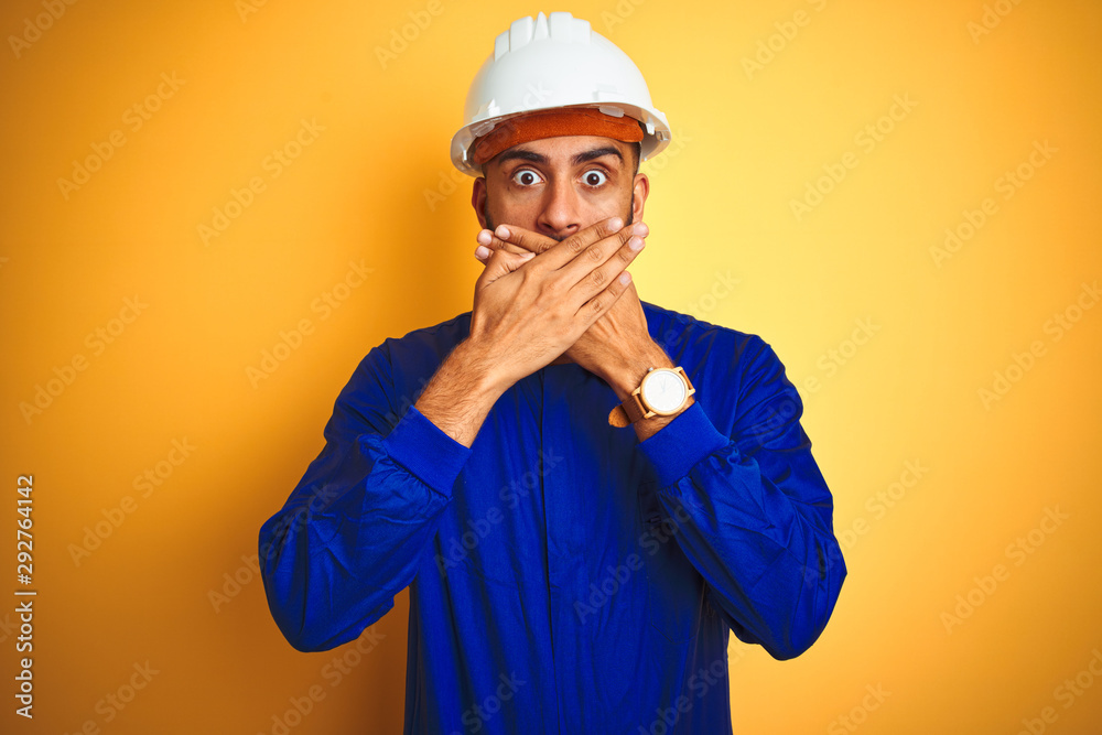 Handsome indian worker man wearing uniform and helmet over isolated yellow background shocked covering mouth with hands for mistake. Secret concept. Handsome indian worker man wearing uniform and helmet over isolated yellow background shocked covering mouth with hands for mistake. Secret concept.