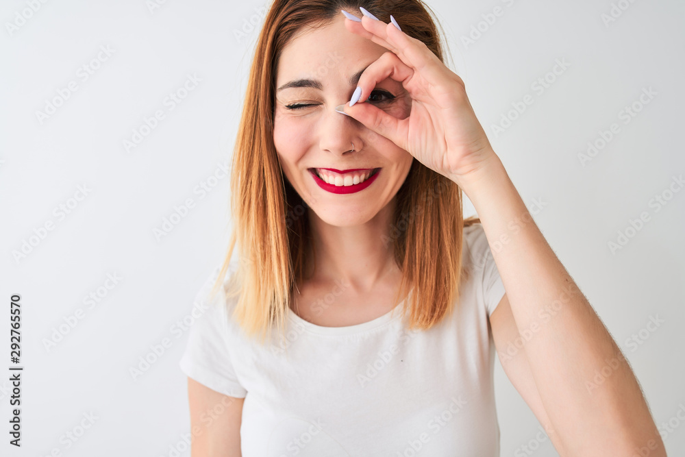 Beautiful redhead woman wearing casual t-shirt standing over isolated white background with happy face smiling doing ok sign with hand on eye looking through fingers Beautiful redhead woman wearing casual t-shirt standing over isolated white background with happy face smiling doing ok sign with hand on eye looking through fingers