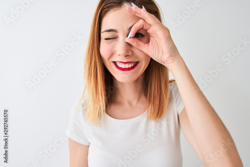 Beautiful redhead woman wearing casual t-shirt standing over isolated white background with happy face smiling doing ok sign with hand on eye looking through fingers