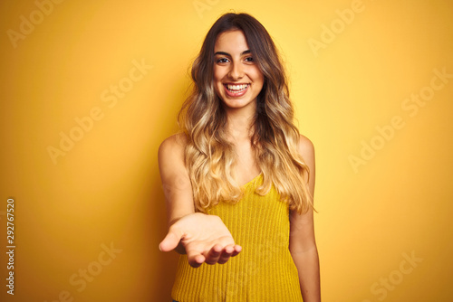 Young beautiful woman wearing t-shirt over yellow isolated background smiling cheerful offering palm hand giving assistance and acceptance.
