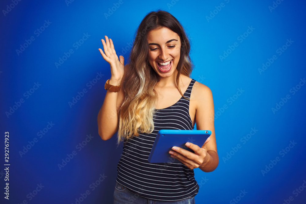 Fototapeta premium Young beautiful woman using touchpad tablet over blue isolated background very happy and excited, winner expression celebrating victory screaming with big smile and raised hands