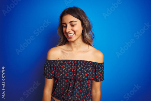 Young beautiful woman wearing floral t-shirt over blue isolated background looking away to side with smile on face, natural expression. Laughing confident.