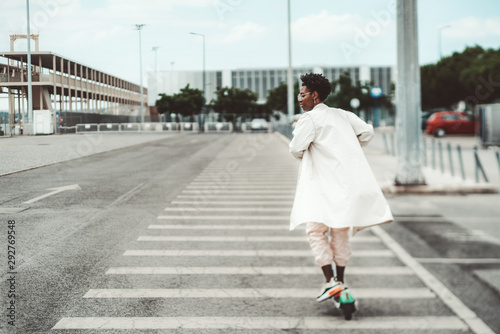 True tilt-shift shot of a young African female in glasses and a white fluttering cloak riding an e-scooter on a wide road with marking on it on a cloudy day