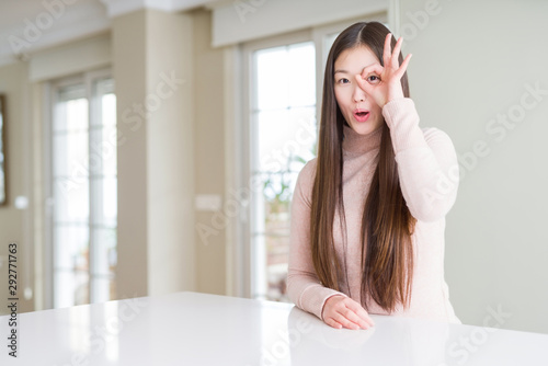Beautiful Asian woman wearing casual sweater on white table doing ok gesture shocked with surprised face  eye looking through fingers. Unbelieving expression.