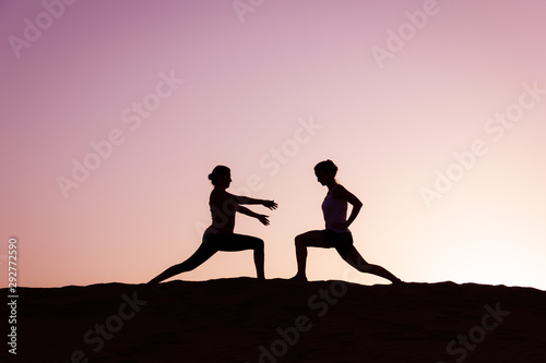 Fototapeta Naklejka Na Ścianę i Meble -  Silhouette of yoga teacher showing warrior pose to student at colorful sunset on Maspalomas sand dunes in Gran Canaria island, Spain