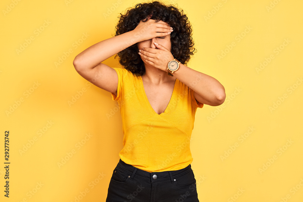 Young arab woman with curly hair wearing t-shirt standing over isolated yellow background Covering eyes and mouth with hands, surprised and shocked. Hiding emotion Young arab woman with curly hair wearing t-shirt standing over isolated yellow background Covering eyes and mouth with hands, surprised and shocked. Hiding emotion