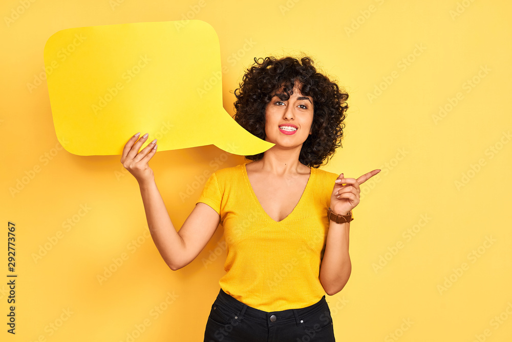 Young arab woman with curly hair holding speech bubble over isolated yellow background very happy pointing with hand and finger to the side Young arab woman with curly hair holding speech bubble over isolated yellow background very happy pointing with hand and finger to the side