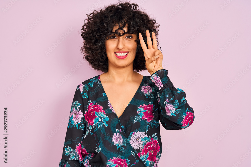 Young arab woman with curly hair wearing floral dress over isolated pink background showing and pointing up with fingers number three while smiling confident and happy. Young arab woman with curly hair wearing floral dress over isolated pink background showing and pointing up with fingers number three while smiling confident and happy.