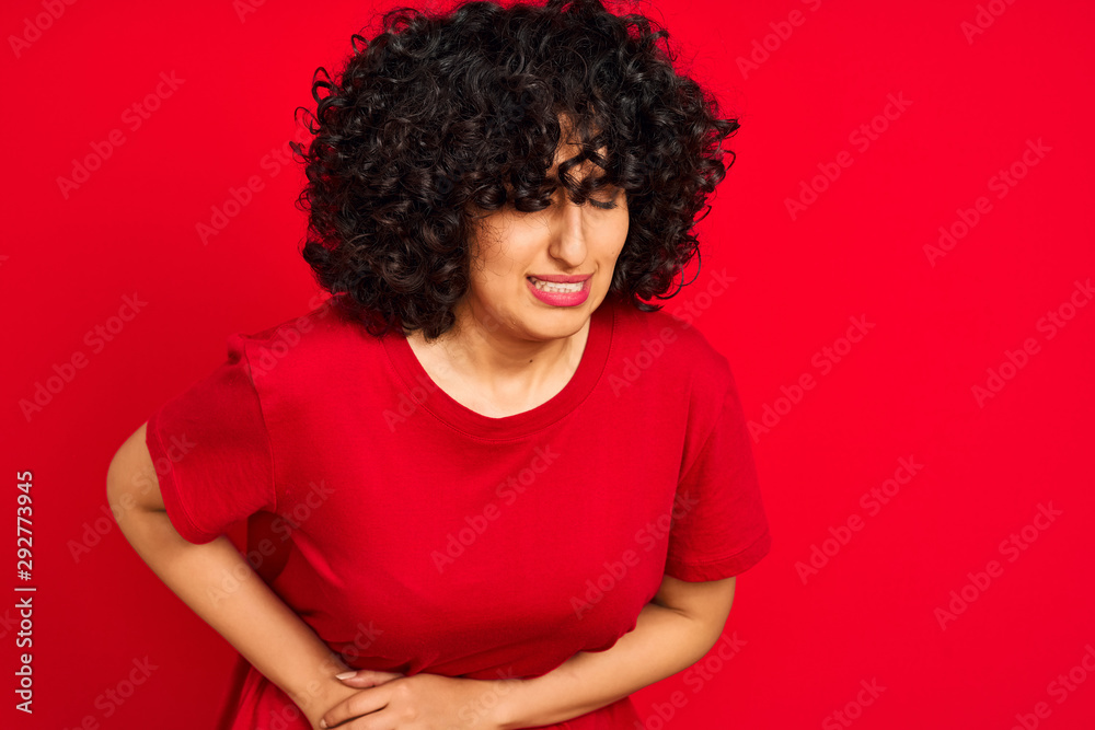 Young arab woman with curly hair wearing casual t-shirt over isolated red background with hand on stomach because nausea, painful disease feeling unwell. Ache concept. Young arab woman with curly hair wearing casual t-shirt over isolated red background with hand on stomach because nausea, painful disease feeling unwell. Ache concept.