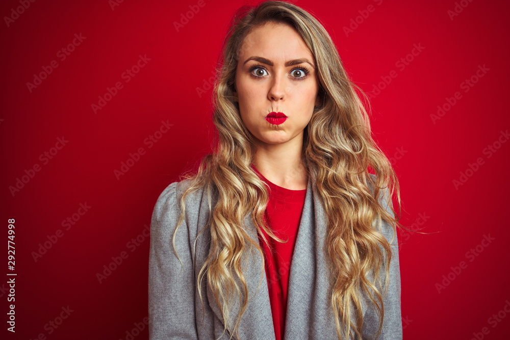 Young beautiful business woman wearing elegant jacket standing over red isolated background puffing cheeks with funny face. Mouth inflated with air, crazy expression.
