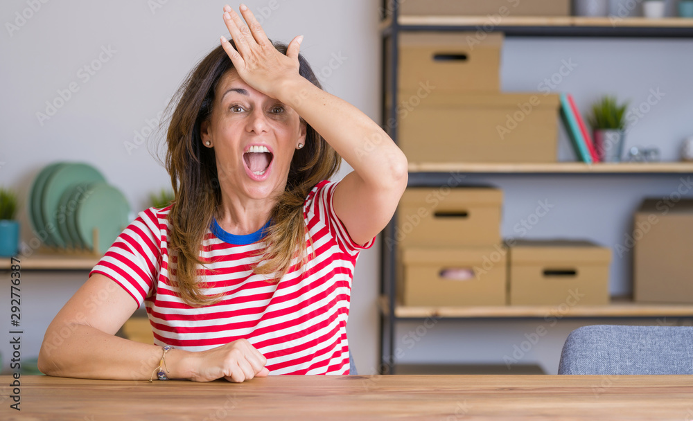 Middle age senior woman sitting at the table at home surprised with hand on head for mistake, remember error. Forgot, bad memory concept. Middle age senior woman sitting at the table at home surprised with hand on head for mistake, remember error. Forgot, bad memory concept.