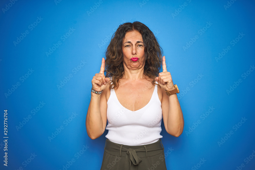 Fototapeta premium Middle age senior woman with curly hair standing over blue isolated background Pointing up looking sad and upset, indicating direction with fingers, unhappy and depressed.