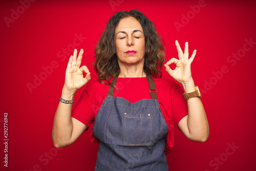 Middle age senior woman wearing apron uniform over red isolated background relaxed and smiling with eyes closed doing meditation gesture with fingers. Yoga concept. © Krakenimages.com