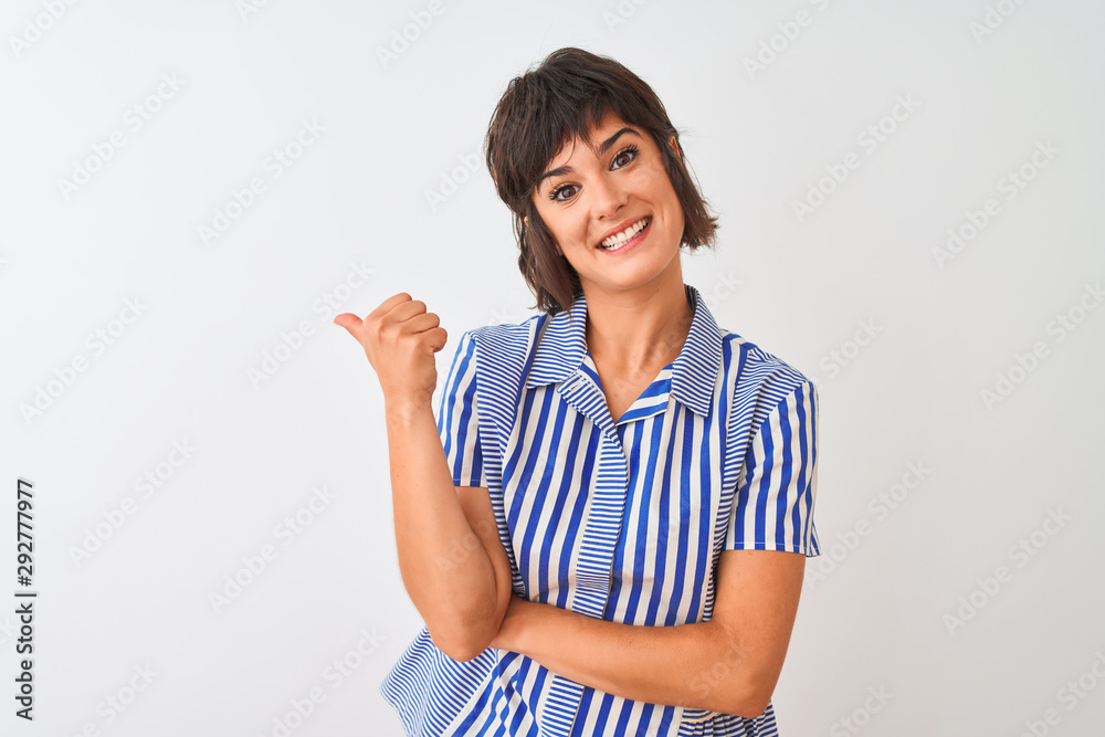 Young beautiful woman wearing blue striped shirt standing over isolated white background smiling with happy face looking and pointing to the side with thumb up. Young beautiful woman wearing blue striped shirt standing over isolated white background smiling with happy face looking and pointing to the side with thumb up.