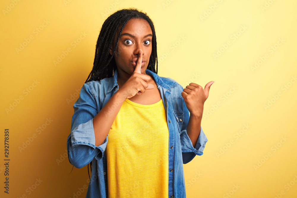 Young african american woman wearing denim shirt standing over isolated yellow background asking to be quiet with finger on lips pointing with hand to the side. Silence and secret concept. Young african american woman wearing denim shirt standing over isolated yellow background asking to be quiet with finger on lips pointing with hand to the side. Silence and secret concept.