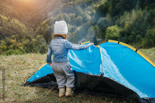 A pretty child prepares his blue tent of outdoors looking at mountain view. Camping girl in scenic landscape enjoying vacation travel adventure nature. Travel  people and healthy lifestyle concept