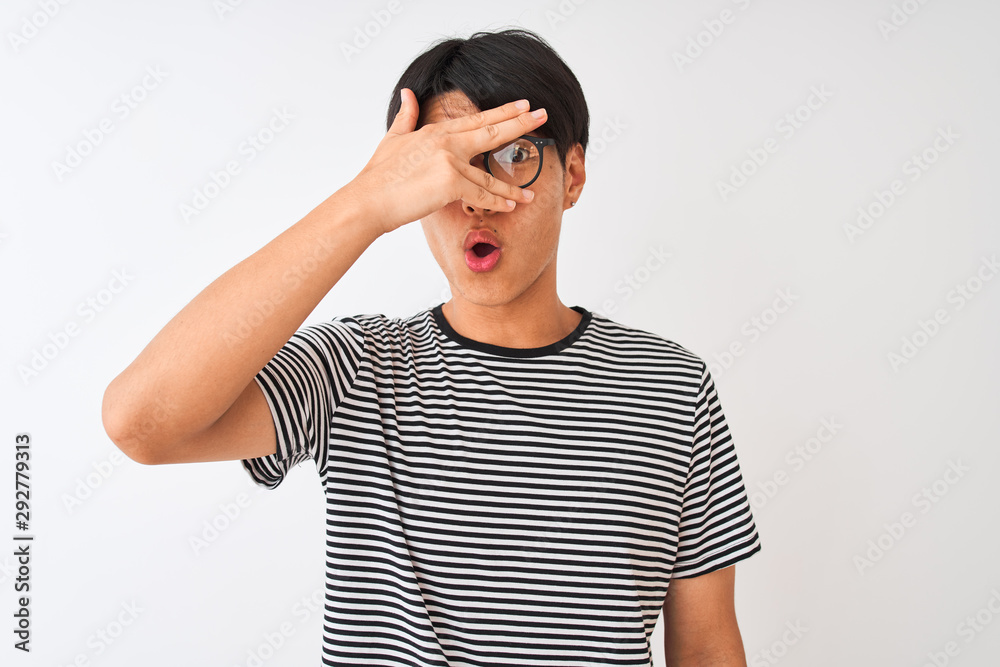 Chinese man wearing glasses and navy striped t-shirt standing over isolated white background peeking in shock covering face and eyes with hand, looking through fingers with embarrassed expression. Chinese man wearing glasses and navy striped t-shirt standing over isolated white background peeking in shock covering face and eyes with hand, looking through fingers with embarrassed expression.