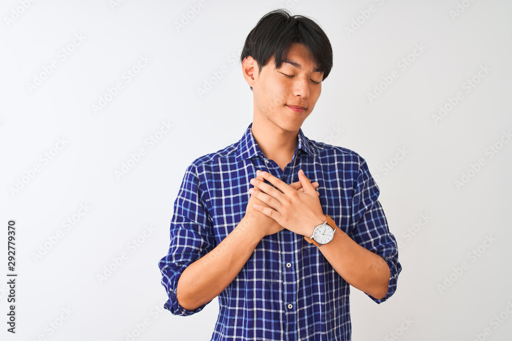 Young chinese man wearing casual blue shirt standing over isolated white background smiling with hands on chest with closed eyes and grateful gesture on face. Health concept. Young chinese man wearing casual blue shirt standing over isolated white background smiling with hands on chest with closed eyes and grateful gesture on face. Health concept.