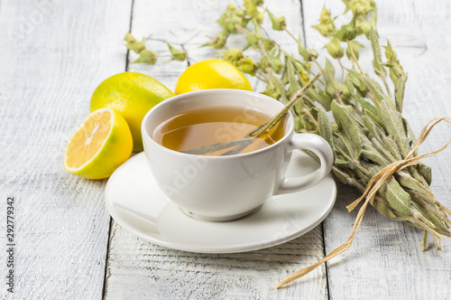 White cup of herbal sage tea with dried sage leaves and lemon on white wooden rustic background. Herbal tea hot drink concept,  Salvia officinalis