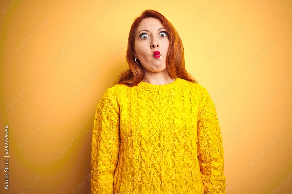 Beautiful redhead woman wearing winter sweater standing over isolated yellow background making fish face with lips, crazy and comical gesture. Funny expression. Beautiful redhead woman wearing winter sweater standing over isolated yellow background making fish face with lips, crazy and comical gesture. Funny expression.
