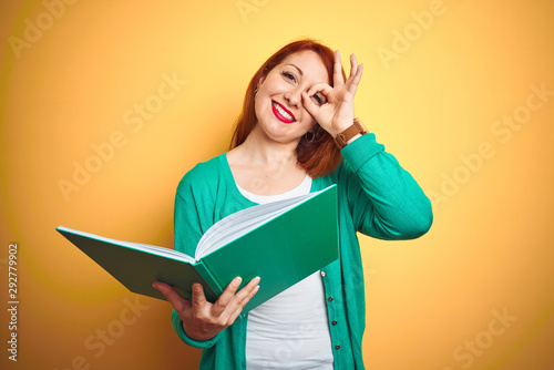 Young redhead student woman reading green book over yellow isolated background with happy face smiling doing ok sign with hand on eye looking through fingers