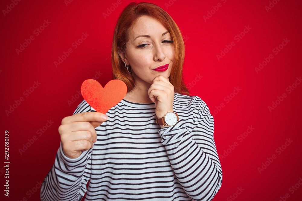 Beautiful redhead romantic woman holding heart standing over isolated red background serious face thinking about question, very confused idea Beautiful redhead romantic woman holding heart standing over isolated red background serious face thinking about question, very confused idea