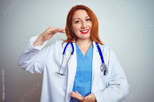 Young redhead doctor woman using stethoscope over white isolated background gesturing with hands showing big and large size sign  measure symbol. Smiling looking at the camera. Measuring concept.