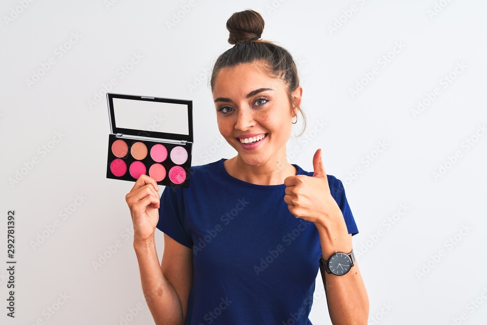 Young beautiful woman holding makeup standing over isolated white background happy with big smile doing ok sign, thumb up with fingers, excellent sign Young beautiful woman holding makeup standing over isolated white background happy with big smile doing ok sign, thumb up with fingers, excellent sign