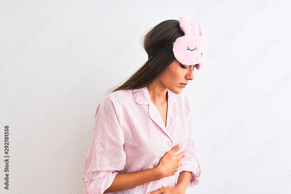 Young beautiful woman wearing sleep mask and pajama over isolated white background with hand on stomach because indigestion, painful illness feeling unwell. Ache concept. Young beautiful woman wearing sleep mask and pajama over isolated white background with hand on stomach because indigestion, painful illness feeling unwell. Ache concept.