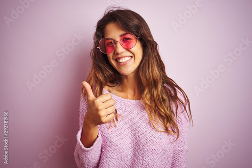 Young beautiful woman wearing fashion sunglasses standing over pink isolated background doing happy thumbs up gesture with hand. Approving expression looking at the camera with showing success.